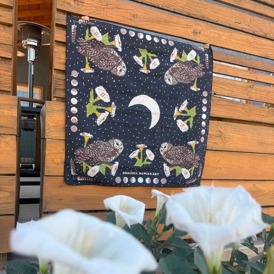 A square black fabric bandana or scarf featuring a moon and owl pattern, hanging on a wooden slat door with white flowers in the foreground.
