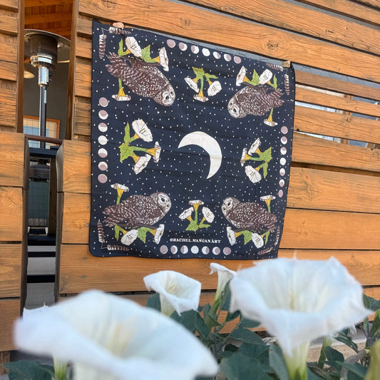A square black fabric bandana or scarf featuring a moon and owl pattern, hanging on a wooden slat door with white flowers in the foreground.