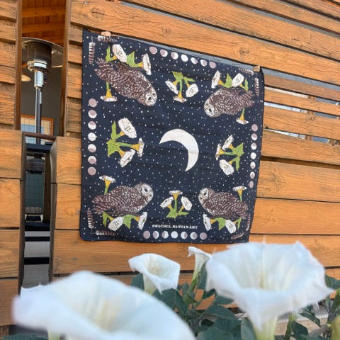 A square dark blue cotton/silk fabric bandana featuring a moon and owl pattern, hanging on a wooden slat door with white datura flowers in the foreground.