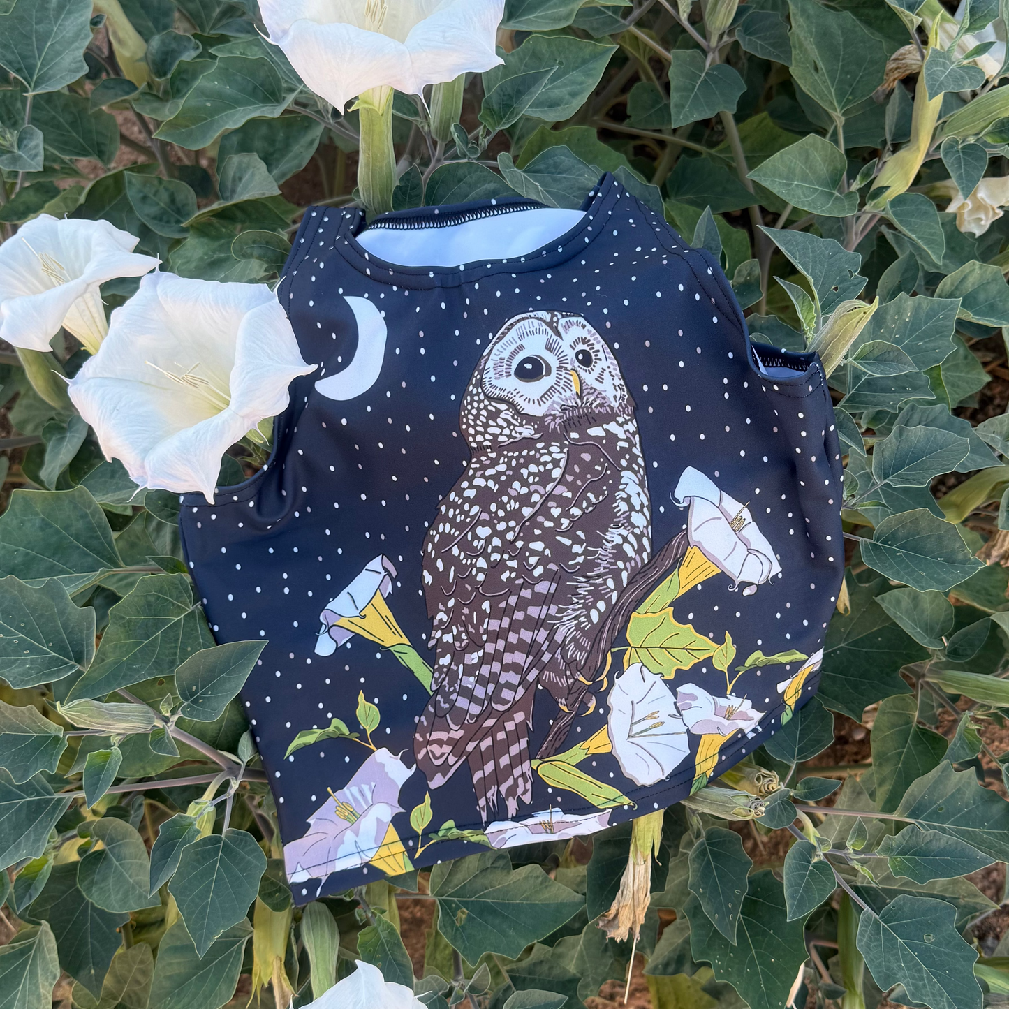 A dark blue crop top featuring a Mexican Spotted Owl, datura and crescent moon, laying on top of a blooming datura bush