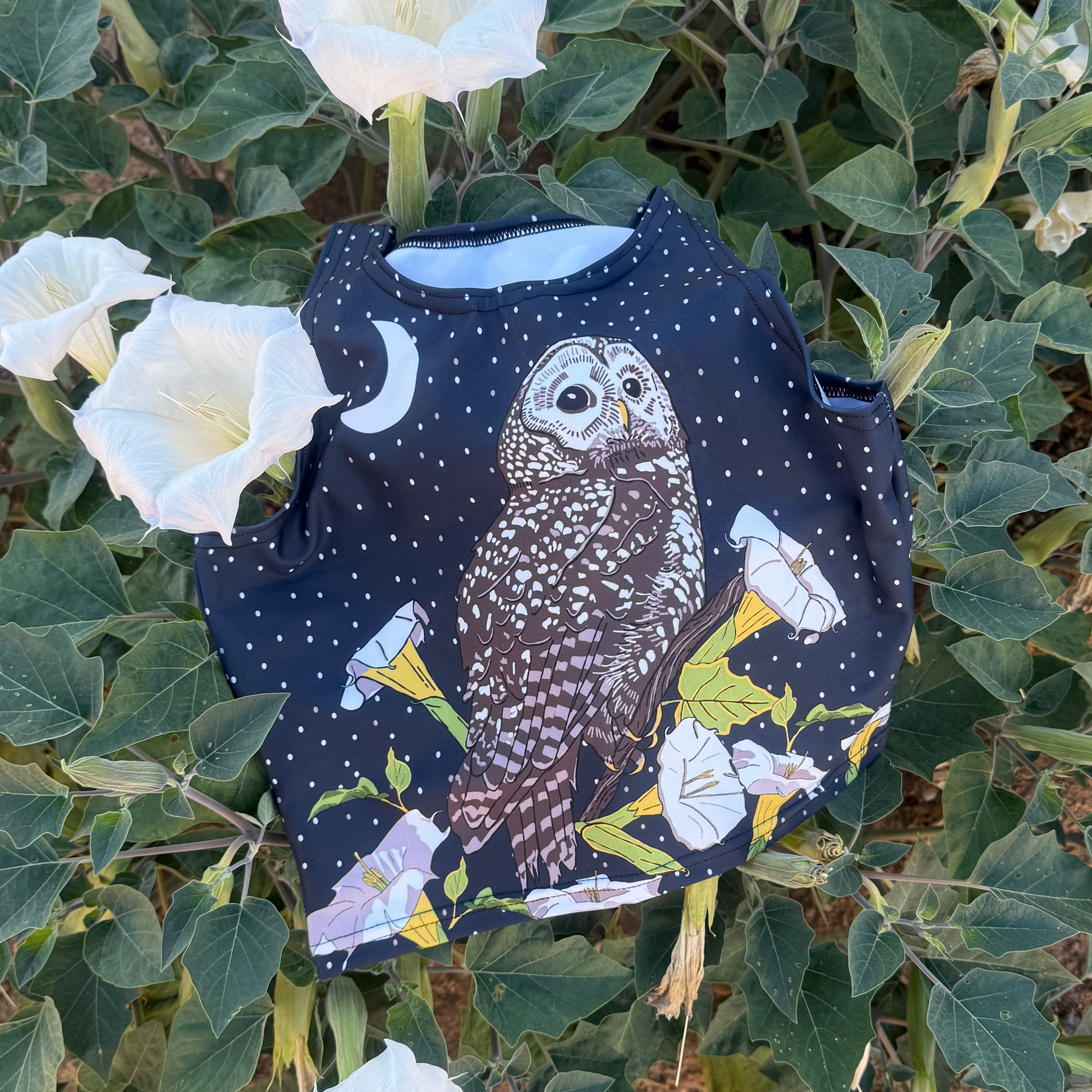 A dark blue crop top featuring a Mexican Spotted Owl, datura and crescent moon, laying on top of a blooming datura bush