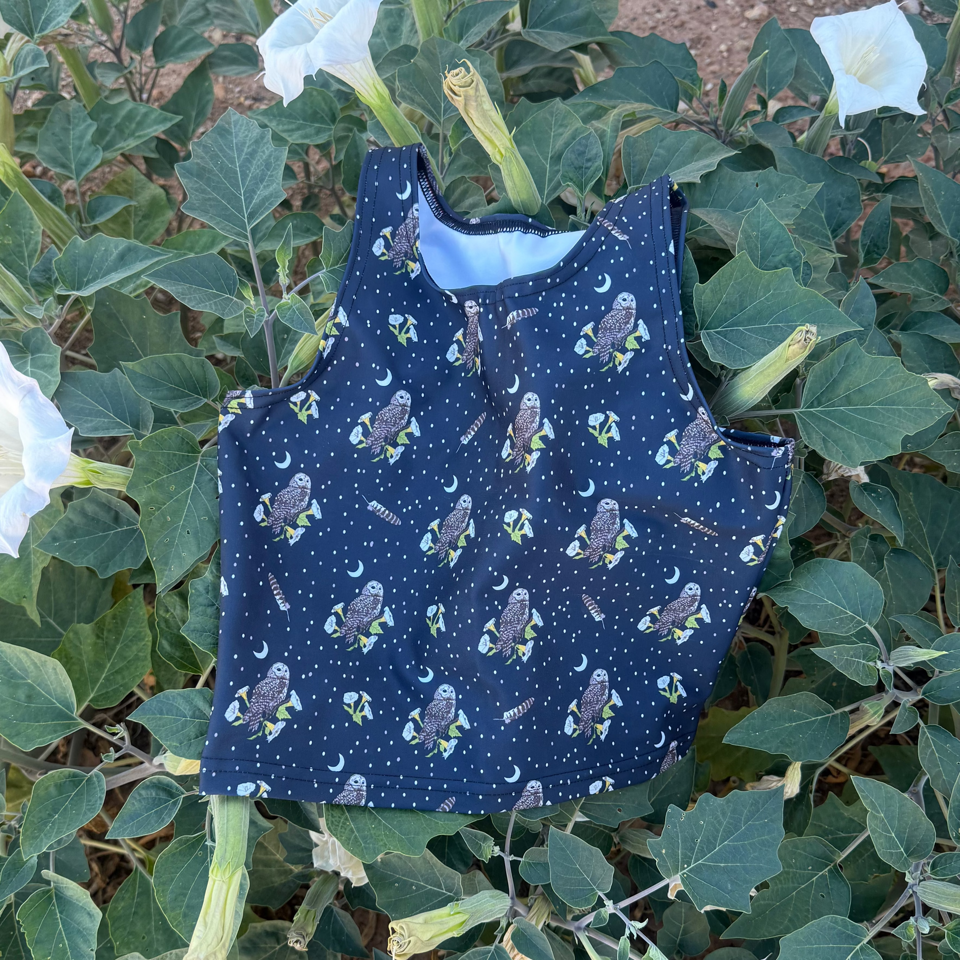 A sleeveless navy blue top with a repeat mexican spotted owl, feather, and datura pattern, laid flat on a flowering datura bush.
