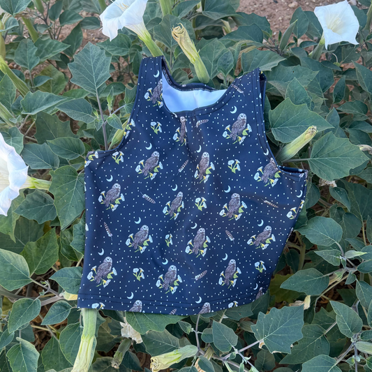 A sleeveless navy blue top with a repeat mexican spotted owl, feather, and datura pattern, laid flat on a flowering datura bush.